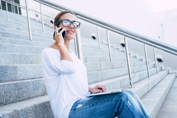 Young businesswoman sitting outdoor with laptop and phone calling and working free in remote smart office - modern people and connection internet job lifestyle - female speaking at cell