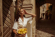 Summer outdoor portrait of fashionable curly woman wearing white outfit posing with lemons in sunny street of European city. Fashion, lifestyle conception. Copy, empty space for text