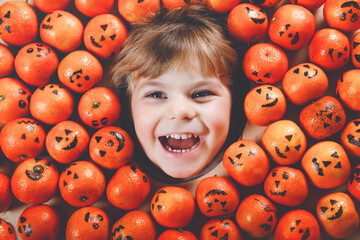 Funny portrait of adorable toddler girl with orange tangerine looking as small jack-o-lantern pumpkins. Happy smiling child celebration Halloween festival or party. Making postcards for friends.