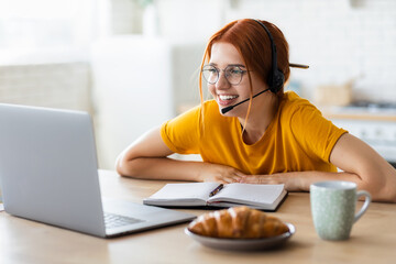 Online learning, portrait of red-haired young caucasian smiling student woman in headset with microphone involved online conference, watching training webinar looking at laptop screen