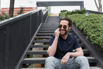 happy man in sunglasses and polo shirt talking on smartphone outside.