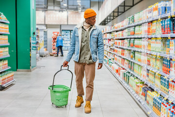 Wall Mural - African-American guy with green plastic basket walks looking at shelves with different goods in modern supermarket department