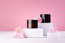 Cosmetic Mockup -  Jars For Cream Of Amber Glass On White Podiums With Pink Spring Flowers. Template For Branding Identity For Cosmetics Produce.