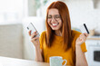 © kucherav - Young cheerful caucasian girl doing online shopping at home using mobile phone and credit card, redhead beautiful excited girl feels euphoria from a long-awaited purchase the Internet, smiling