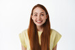 © Cookie Studio - Close up of hopeful pretty girl with long red hair and freckles, looking below camera at tv screen, smiling happy and pleased with results, standing over white background