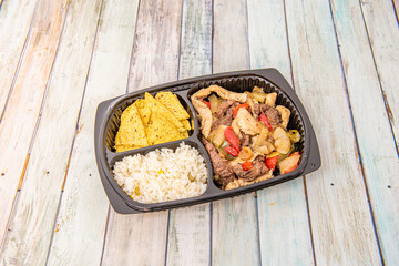 Black home delivery tray with mixed chicken and beef fajitas with corn nachos and white rice on wooden background