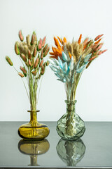 two pretty glass vases with spikes of dried plants impregnated with bold colors on varnished gray metal table