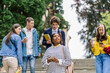 © Iryna - Positive brooding attractive african american female student thinking and looking upward standing on stairs while people, fellow students are walking around her.