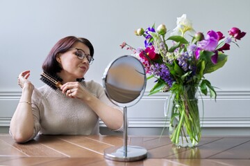 Wall Mural - Middle aged woman styling her hair with round comb in front of makeup mirror at home