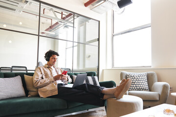 young man online with mobile phone and headphones relaxing in seating area of open plan office