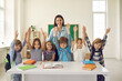 © Studio Romantic - Portrait of cheerful female teacher and little students with bags standing together near table in modern classroom. Happy school kids and their tutor smiling, raising hands and looking at camera
