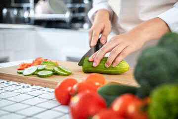 Wall Mural - Close up of asian housewife wearing apron and using knife to slice cucumber and tomato on chopping board