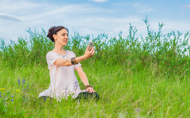 Wall Mural - A beautiful young woman sits on a green lawn in a lotus position, holds a phone in her hand and looks at it. Outdoor recreation.