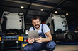 © Dusan Petkovic - Smiling tattooed bearded blue collar worker in overalls using tablet to check on delivery while crouching in garage of import and export firm. In background are trucks.