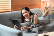 © Prathankarnpap - Happy young woman holding digital table and smiling to camera while lying on sofa.