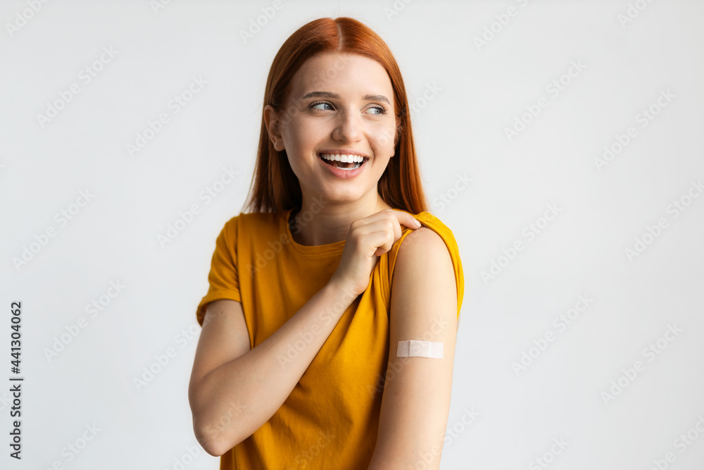 Covid-19 vaccinated caucasian smiling young woman showing arm with plaster, gray background
