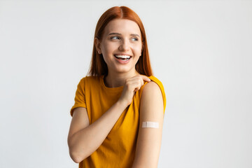 Wall Mural - Covid-19 vaccinated caucasian smiling young woman showing arm with plaster, gray background