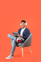 Canvas Print - Young African-American guy with tablet computer listening to music while sitting in armchair on color background