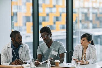 Multi-ethnic group of doctors discussing case while sitting at meeting table during medical council, copy space