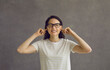 I can't hear you. Head shot of young woman in glasses closing eyes and plugging ears with fingers to ignore loud noise, unwanted opinion, dumb stupid comment or useless unsolicited uninvited criticism