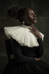 Wall Mural - Close-up portrait of medieval African young woman in black vintage dress with big white collar posing isolated on dark green background.