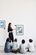 © Seventyfour - Vertical back view at diverse group of children sitting on floor in modern art gallery and looking at paintings, copy space