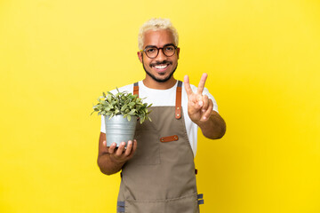 Wall Mural - Young Colombian man holding a plant isolated on yellow background smiling and showing victory sign