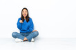 © luismolinero - Young mixed race woman sitting on the floor isolated on white background surprised and pointing front