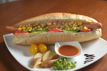 Long hot dog with two sausages, with green peas and corn, with slices of tomatoes and cucumbers, on a white plate with sauce, potatoes and cherry tomatoes