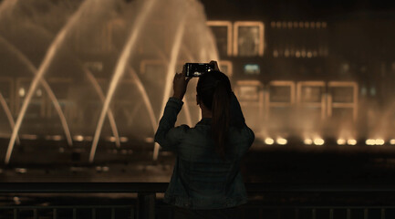 Rear view of a charming tourist girl taking photo or video of fountains