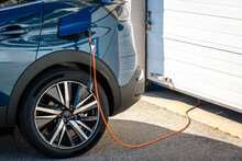 A Hybrid Electric Car Parked In Front Of The Home Garage Door Is Plugged In With A Charging Cable To Recharge The Battery.