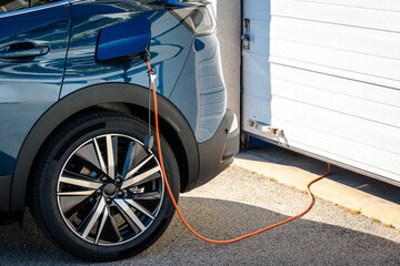 A hybrid electric car parked in front of the home garage door is plugged in with a charging cable to recharge the battery.