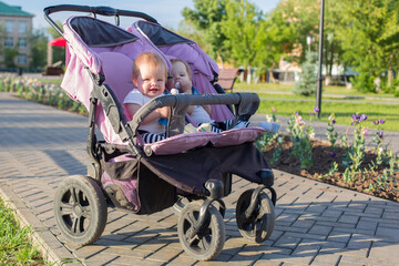 Twins on a walk in a stroller eat fruit puree in city park.