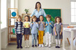 © Studio Romantic - Teacher and children on first school day. Full body length portrait of happy young teacher and little group of elementary students in casual wear standing in classroom, looking at camera and smiling