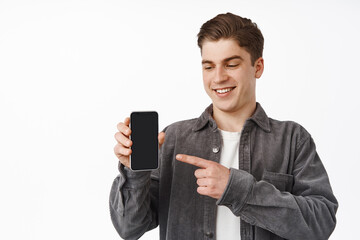 Wall Mural - Close up of cheerful caucasian guy pointing finger at smartphone, showing mobile phone screen app, interface application, standing against white background