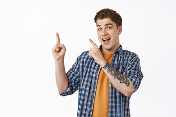 Wall Mural - Portrait of smiling young man pointing fingers at upper left corner, invite to click link, showing shopping discounts, give info, advertise something, standing over white background