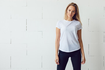 Blonde girl in a white blank t-shirt stands against the background of a white wall.