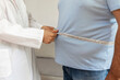 © Jelena Stanojkovic - A nutritionist doctor measures the body of a male patient with a measuring tape on adipose tissue and excess weight. Overweight obesity man seeing doctor for treatment of his illness and weight loss.