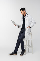 Wall Mural - Side view of arabian doctor using laptop near chair on grey background