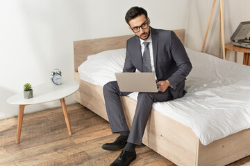 Wall Mural - Businessman in eyeglasses holding laptop on bed near alarm clock on bedside table