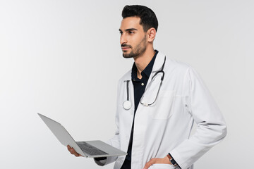 Canvas Print - Young muslim doctor holding laptop and looking away isolated on grey