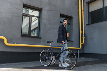 Wall Mural - Arabian businessman with paper cup looking at camera near bike outdoors