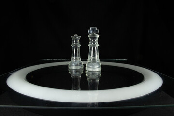 clear queen and king chess pieces isolated on a black background with a white light circle below