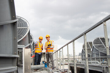 Asian engineer refrigeration plant  factory. male mechanic wearing helmet and Reflective Safety Vest uses laptop, tablet, equipment with sets of cooling towers conditioning systems background.