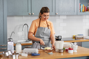 Wall Mural - culinary, baking and people concept - happy smiling young woman cooking food on kitchen at home cutting butter with knife