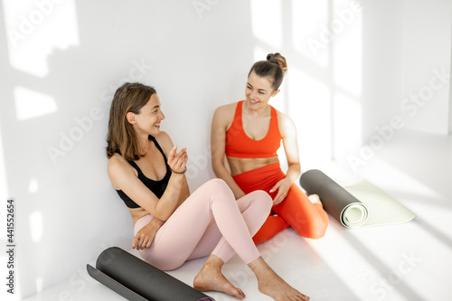 Two female friends in sportswear sitting together with yoga mats and having talk during a yoga training break at gym on the white background
