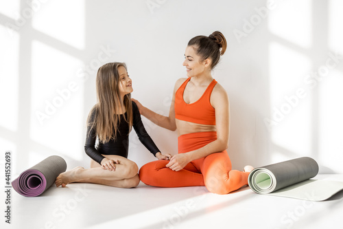 Sports mom and little daughter sitting together with yoga mats and have a close conversation during sports indoors. Close relationships with mother and sports in childhood