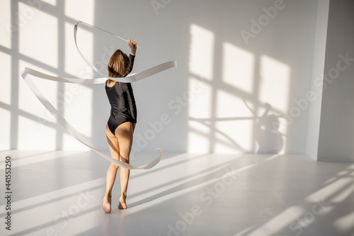 Little girl practising rhythmic gymnastics with a gymnastic tape at white sunny dance room. Wide view with copy space and shadows on the background