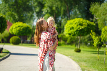 A charming girl in a light summer sundress walks in a green park with her little daughter, holding her in her arms. Enjoys warm sunny summer days