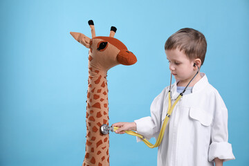 Sticker - Cute little boy in pediatrician's uniform playing with stethoscope and toy giraffe on light blue background